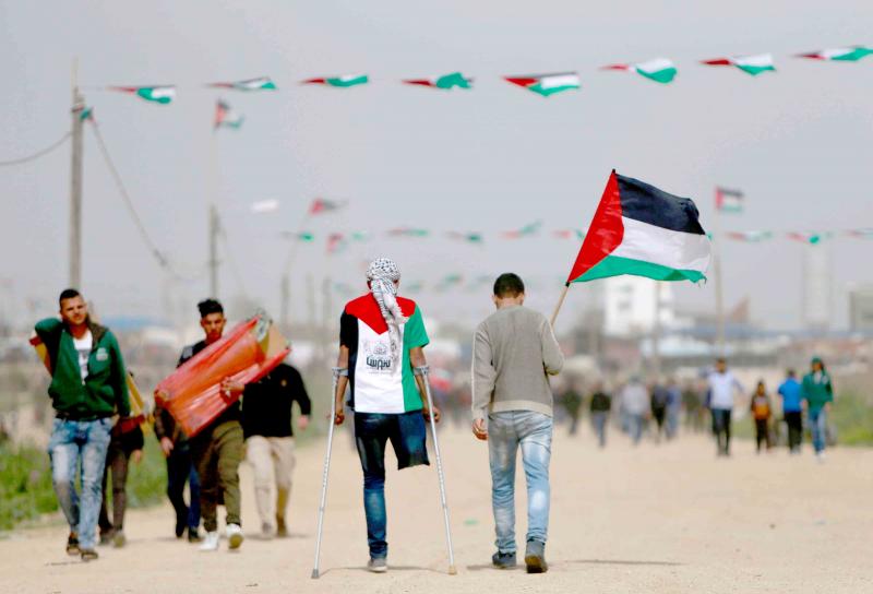 A Palestinian man wearing a t-shirt depicting the colours of the Palestinian flag walks on crutches with another holding a national flag as they head towards a demonstration near the border with Israel, March 30