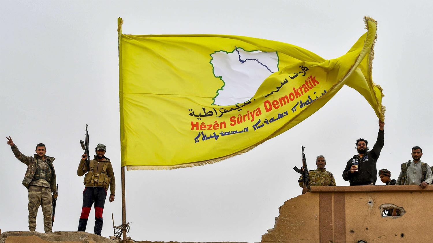 Fighters of the Syrian Democratic Forces (SDF) flash the victory gesture next to their unfurled flag atop a roof at a position in the village of Baghouz 