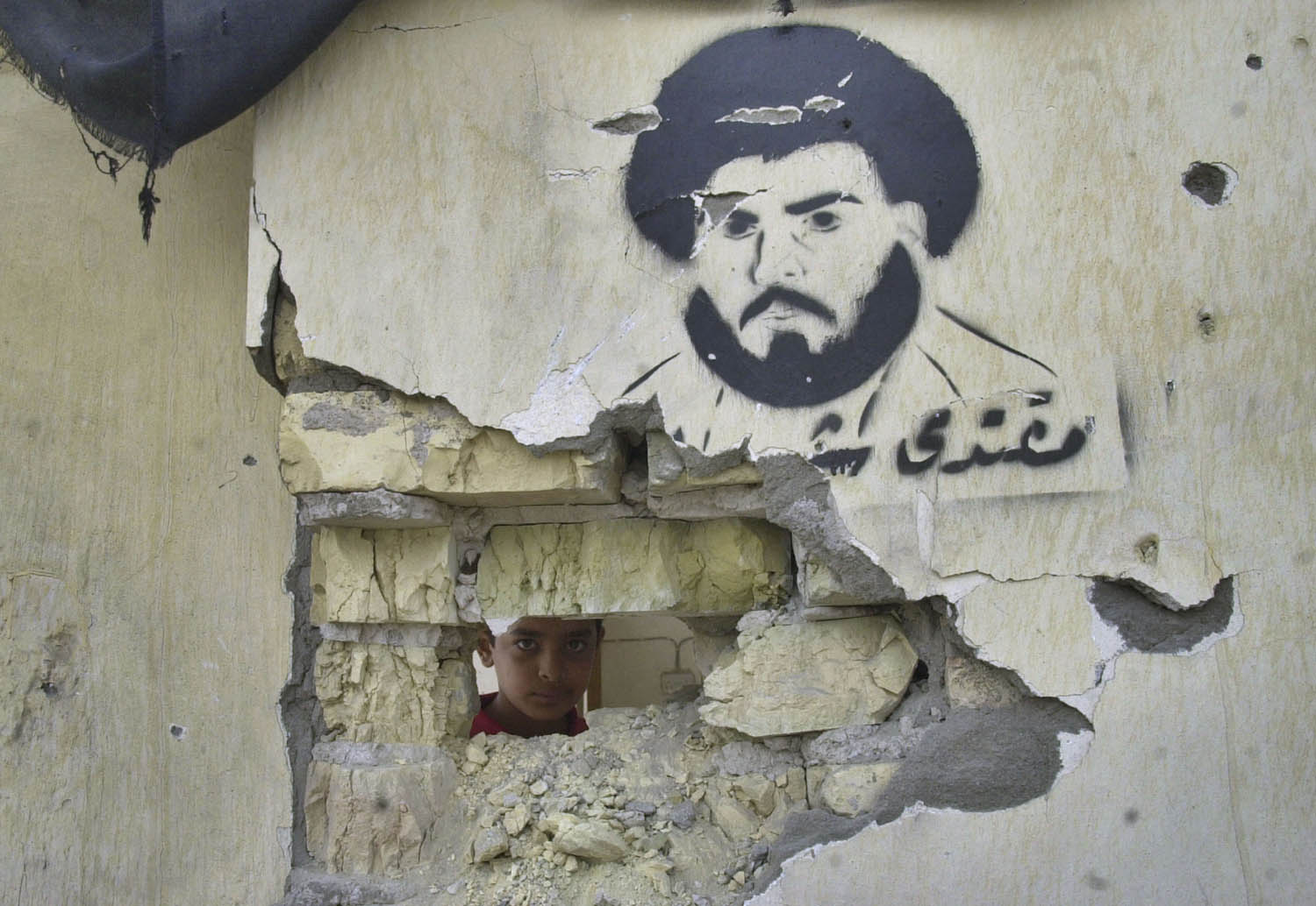 طفل ينظر من فتحة في حائط في مدينة الصدر يحمل صورة لمقتدى الصدر