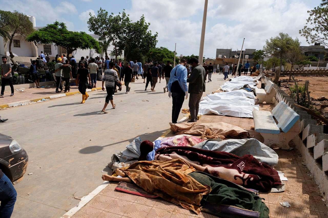 People pass the dead bodies laid on the ground outside the hospital in Derna