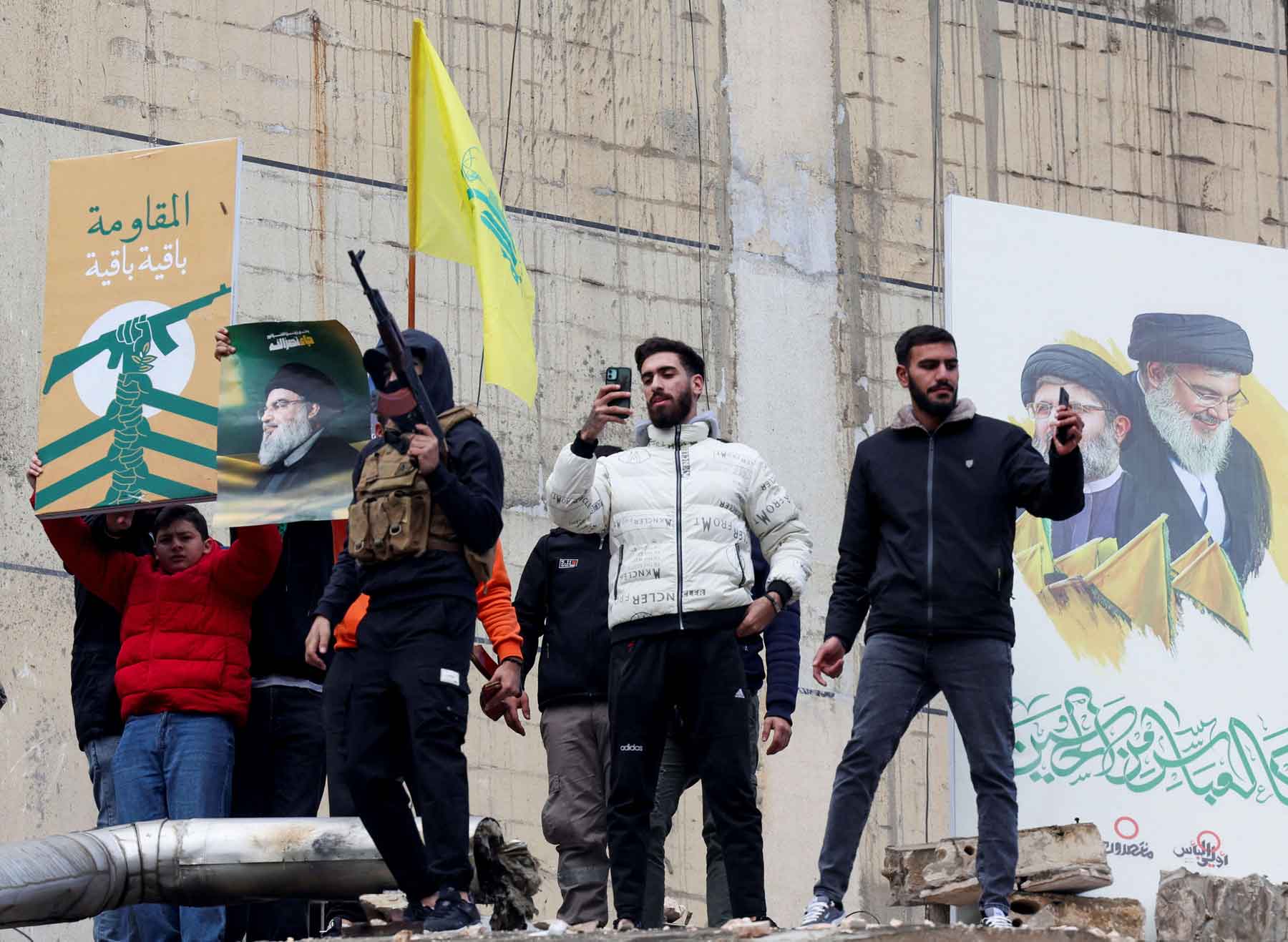 People stand near a poster depicting late Hezbollah leader Sayyed Hassan Nasrallah and late senior Hezbollah official Hashem Safieddine as one of them carries a weapon, in Beirut’s southern suburbs, November 27, 2024. 