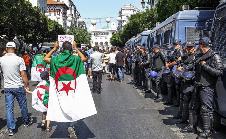 Protesters draped in Algerian national flags walk past riot police members