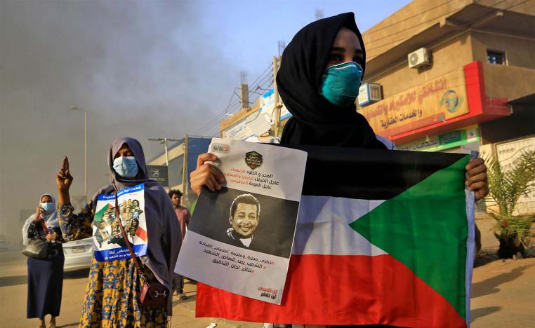 A mask-clad Sudanese woman holds a national flag along with a poster depicting the face of a slain protester