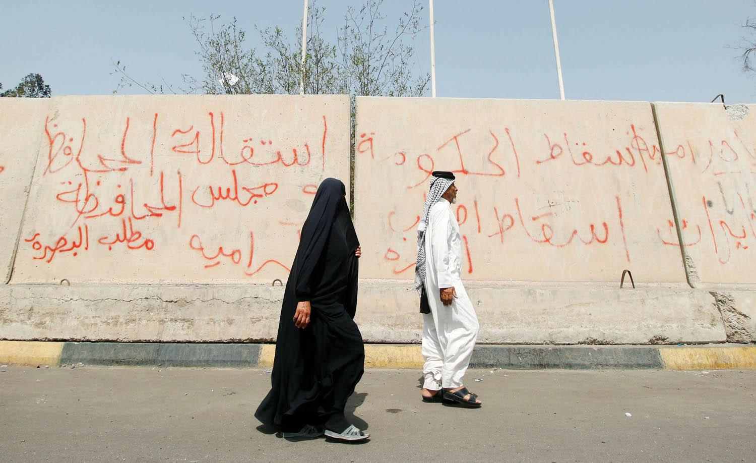 عراقي وعراقية يمران بجانب جدار يحمي منشأة حكومية في البصرة تلطخه الشعارات