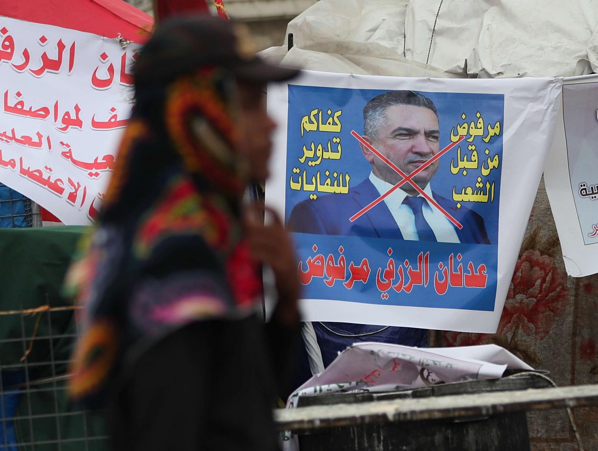 An anti-government protester passes a defaced picture of Iraq's Prime Minister-designate Adnan Al-Zurfi