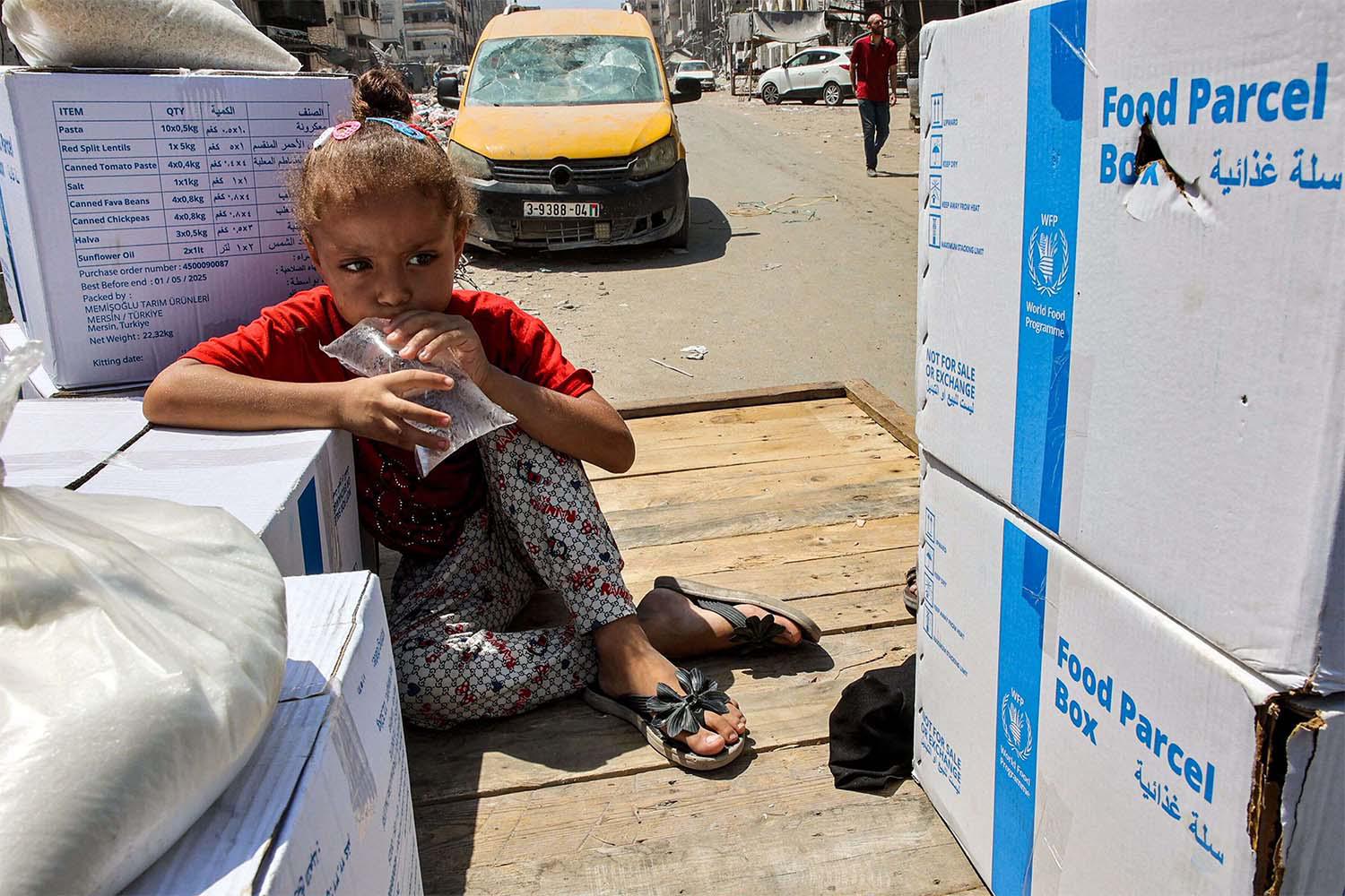 A girl holds a clear plastic bag filled with water as she sits atop a cart next to humanitarian aid packages provided by UNRWA